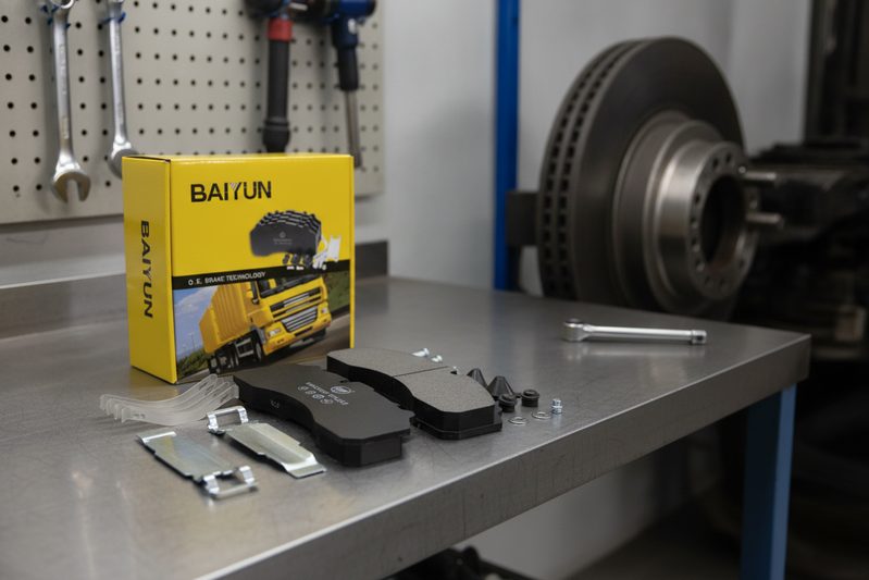 Technician holding BAIYUN commercial vehicle brake pad next to OEM pad, comparing friction material and backing plate for fleet maintenance